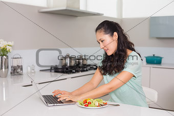 Young woman using laptop while having salad in kitchen