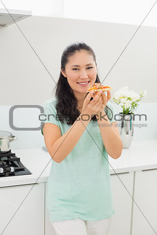 Smiling woman eating a slice of pizza in kitchen