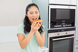 Smiling young woman eating a slice of pizza in kitchen