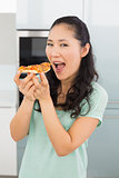 Smiling young woman eating a slice of pizza in kitchen