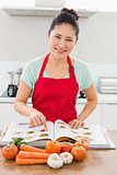 Smiling woman with recipe book and vegetables in kitchen