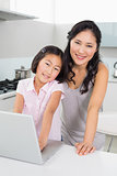 Smiling mother with young daughter using laptop in kitchen