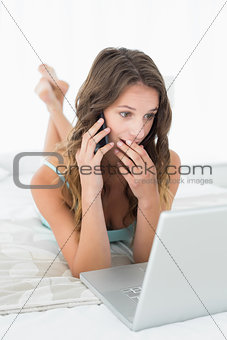 Shocked woman using mobile phone and laptop in bed