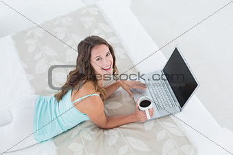 Cheerful young woman using laptop with coffee cup in bed