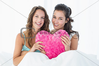 Smiling female friends with heart shaped pillow in bed