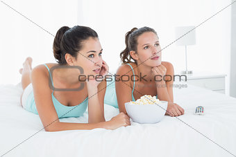 Serious female friends with popcorn bowl lying in bed