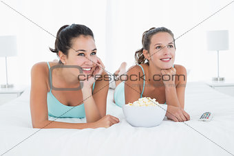 Smiling female friends with popcorn bowl lying in bed