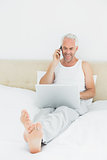 Smiling mature man using cellphone and laptop in bed