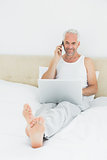 Casual smiling mature man using cellphone and laptop in bed