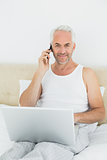 Smiling mature man using cellphone and laptop in bed