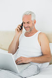 Mature man using cellphone and laptop in bed