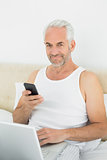 Smiling mature man with cellphone and laptop in bed