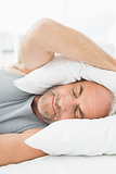 Mature sleepy man covering ears with pillow in bed