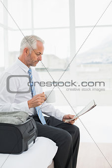 Businessman with coffee cup reading newspaper by luggage at hotel room