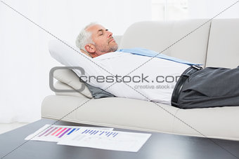 Businessman sleeping on sofa with graphs on table in living room