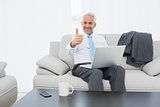 Businessman with laptop gesturing thumbs up at home