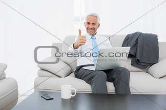 Businessman with laptop gesturing thumbs up at home