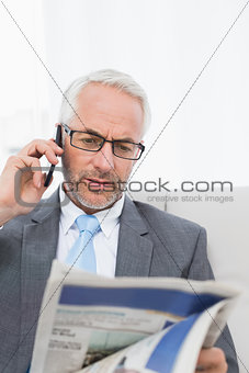Elegant businessman using cellphone while reading newspaper