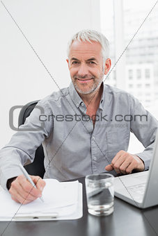 Smiling mature businessman writing notes while using laptop
