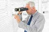 Businessman peeking with binoculars through blinds in office