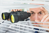 Mature businessman peeking with binoculars through blinds