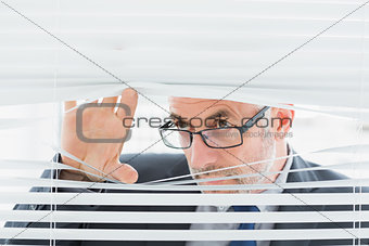 Serious mature businessman peeking through blinds in office