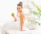 Smiling young woman with apple sitting on massage table