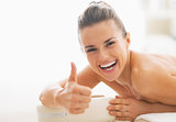 Smiling young woman laying on massage table and showing thumbs u