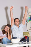 Excited man watching TV with wine and popcorn by bored woman at home