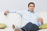 Relaxed man with a drink and remote control sitting on sofa at home
