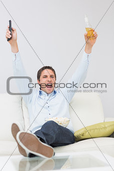 Man with a drink and remote control cheering on sofa