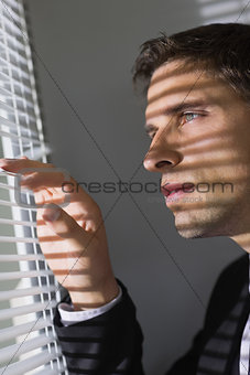 Serious young businessman peeking through blinds in office