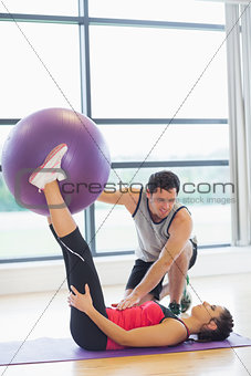Trainer helping young woman with fitness ball at gym