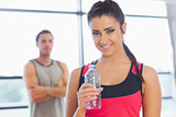 Fit woman holding water bottle with friend in background in exercise room