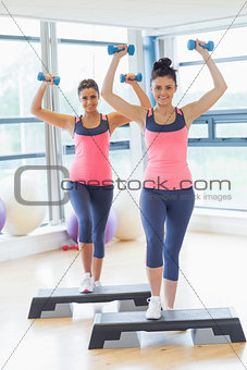 Two women performing step aerobics exercise with dumbbells