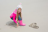 Girl in warm clothing with drawn heart shape on sand at beach