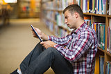 Thoughtful young student sitting on library floor using tablet