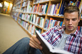 Serious young student sitting on library floor reading book