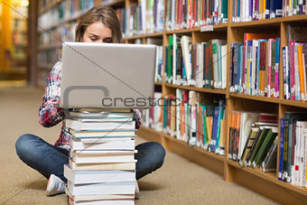Young student sitting on library floor using laptop on pile of books