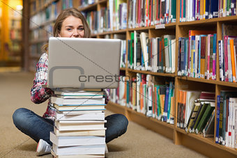 Happy student sitting on library floor using laptop on pile of books