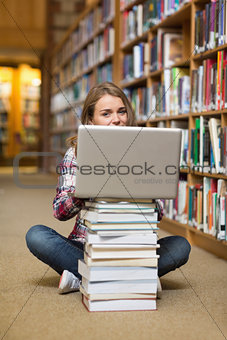 Smiling student sitting on library floor using laptop on pile of books