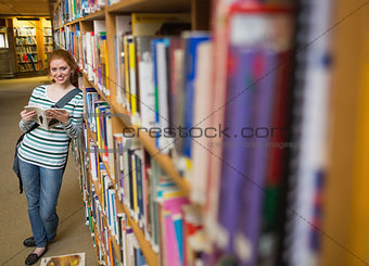 Cheerful student reading book leaning on shelf in library