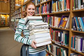 Smiling student holding heavy pile of books standing in library