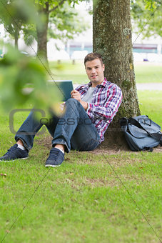 Smiling student using his tablet pc outside leaning on tree