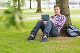 Happy student using his tablet pc outside leaning on tree
