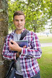 Smiling student leaning on tree holding his tablet pc