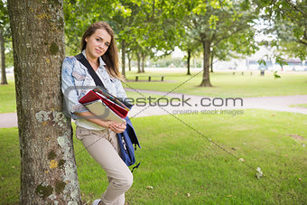 Cheerful student leaning on tree holding her books