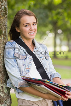 Cheerful young student leaning on tree holding her books