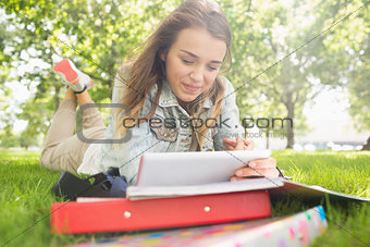 Pretty smiling student lying on the grass studying with her tablet pc