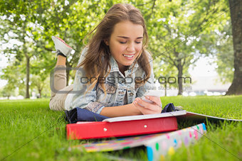Pretty smiling student lying on the grass sending a text